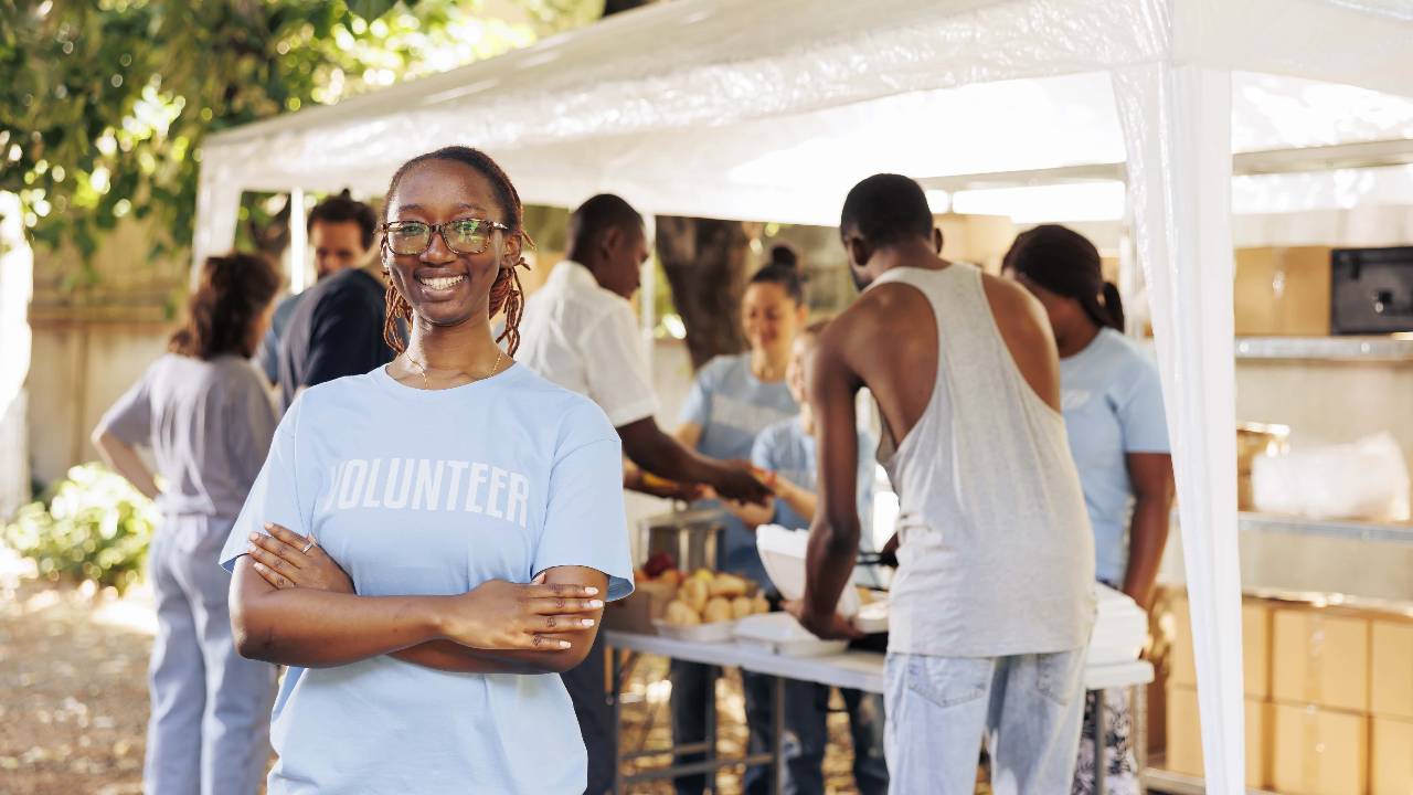 young-black-woman-with-glasses-stands-outdoors-arms-crossed-looking-camera-diverse-group-volunteers-supports-non-profit-program-dedicated-hunger-relief-helping-needy